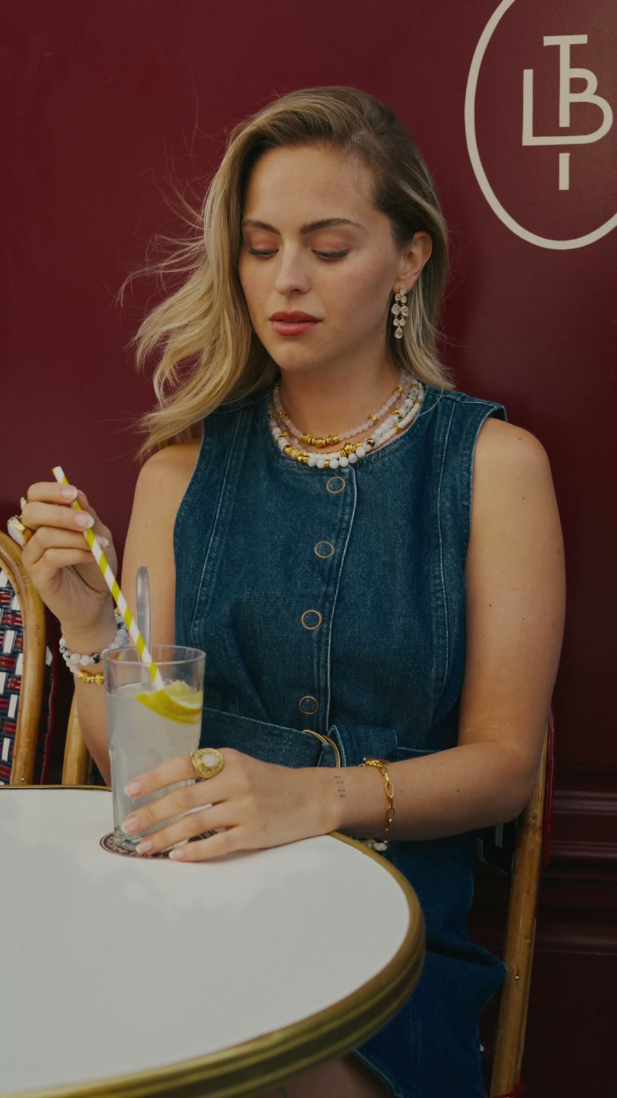 Camille en robe en jean à une terrasse de café parisien, bracelets en pierres naturelles et colliers superposés