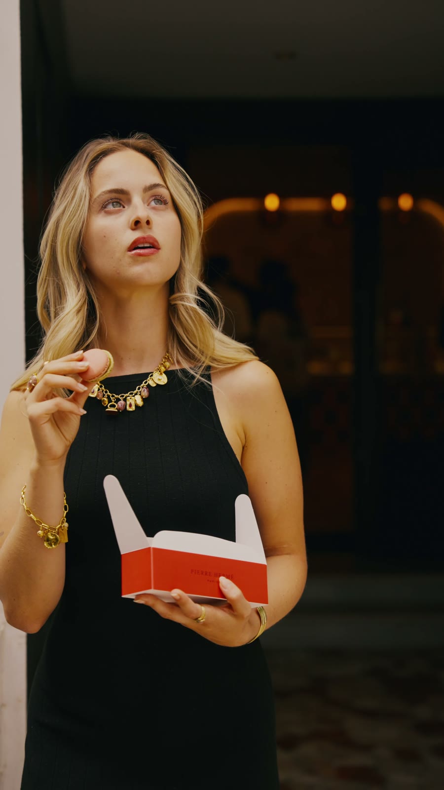 Camille en robe noire tient une boîte de macarons devant la vitrine Pierre Hermé, collier à médailles dorées Eiden