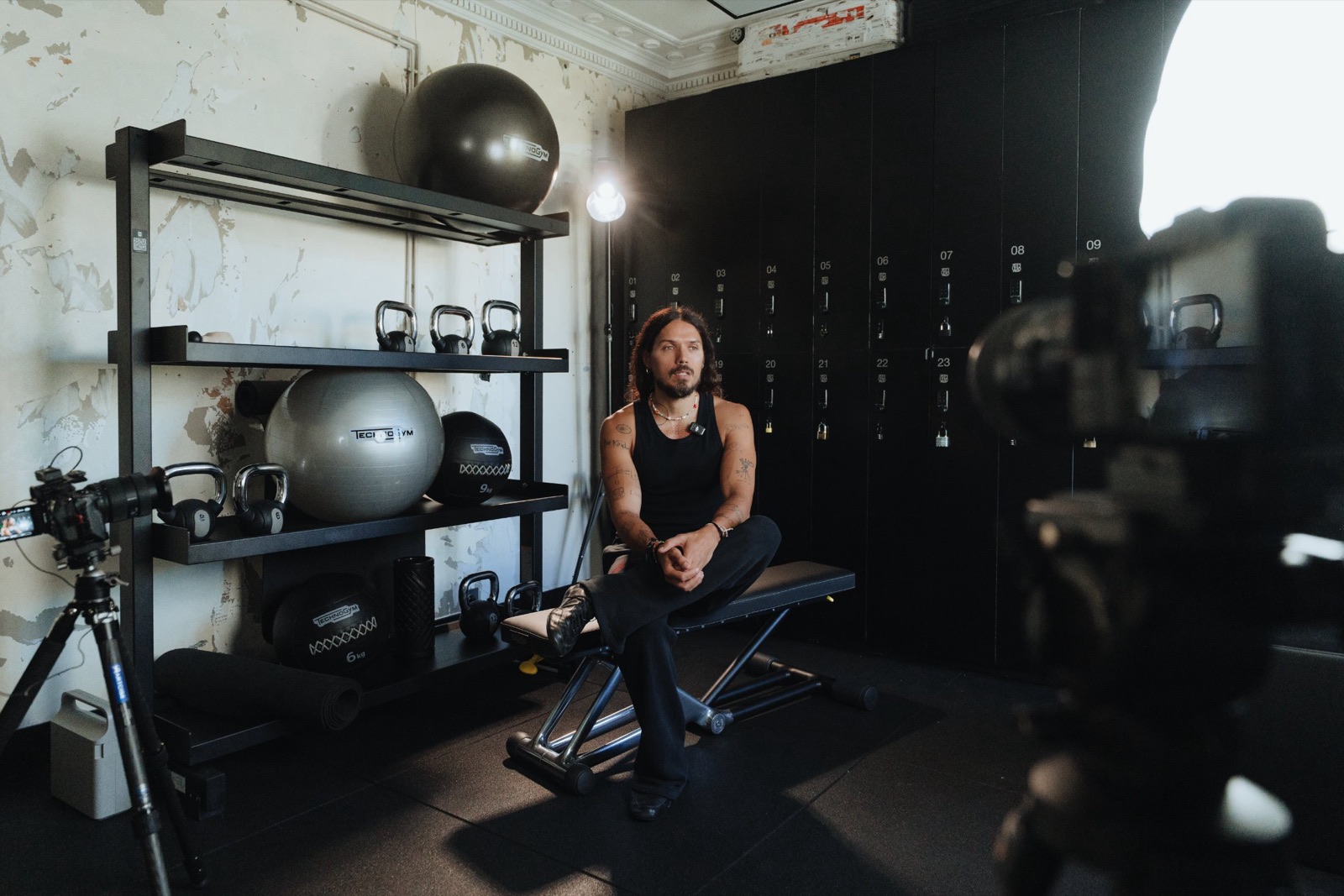 Vue large du plateau : Harry Nuriev assis sur un banc Technogym devant les casiers, caméra au premier plan